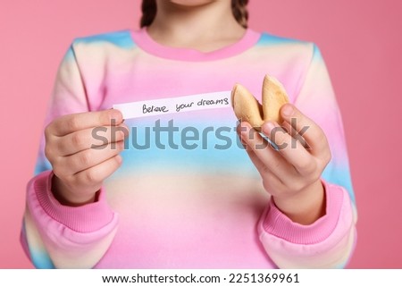 Girl holding tasty fortune cookie and prediction on pink background, closeup