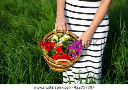Girl holding weaved basket full of roses while standing in high spring grass on sunny meadow. 