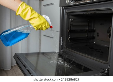 A girl holds a blue spray in her hand against the background of a modern electric oven in the kitchen. Concept for caring and cleaning an electric stove. Care of household appliances.  - Powered by Shutterstock