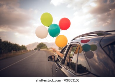 Girl holds colorful balloons out from the window of the car. Freedom, happiness, road trip and celebration concept.
