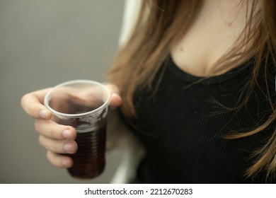 Girl Holds Glass With Drink In Her Hand. Girl Drinking Tea Outside. Black T-shirt And Long Hair. Hot Drink In Plastic Cup.