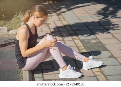 The Girl Holds An Injured Knee Joint In Her Hands. Injury While Running Outdoors.