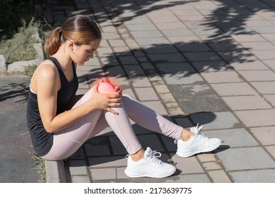 The Girl Holds An Injured Knee Joint In Her Hands. Injury While Running Outdoors.