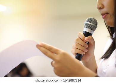 Girl Holds Microphone In Her Hands, Selective Focused Of A Mike Or Mic In A Hand Of A Woman With One Hand In Defocused Mode Holding Paper, Speaking Or Giving Speech On Stage Or In The Training Room