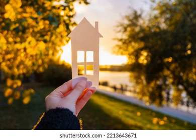 The Girl Holds A Paper House In Her Hand Against The Background Of The Sunset Sky. The Sun Is Setting Over The Horizon. The Concept Of Your Own Home, Personal Space And Mortgage. High Quality Photo