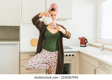 girl in home clothes balances on one leg, standing in home kitchen, with bath towel on her wet hair, pours coffee into cup. Morning coffee in yoga pose at home of teenage girl