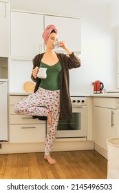 girl in home clothes balances on one leg, standing in home kitchen, with bath towel on wet hair, drinking coffee from cup, holding milk jug in other hand. Morning coffee in yoga pose of teenage girl 
