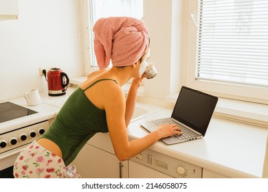 girl in home clothes, tank top with straps, with wound towel on wet hair, view from back, drinks coffee and looks laptop screen, typing with one hand, in home kitchen in morning or on day off