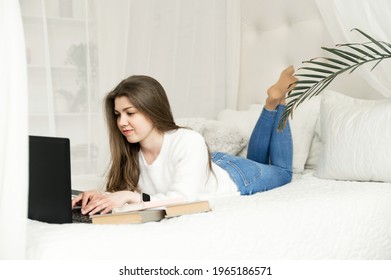 Girl At Home Studying Distance Learning. A Student Sits At A Laptop In Her Bed. Remote Work. Online Education Of A Young Girl