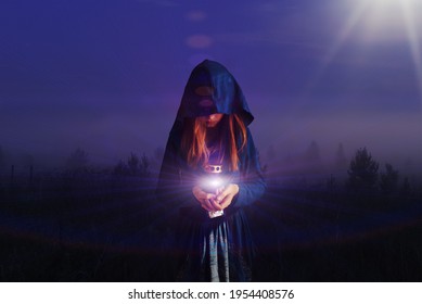 A girl with a hood on her head walks through the night forest and carries a lamp. The light spreads and illuminates the night. The concept of prayer, holiness, mysticism, magic. - Shutterstock ID 1954408576