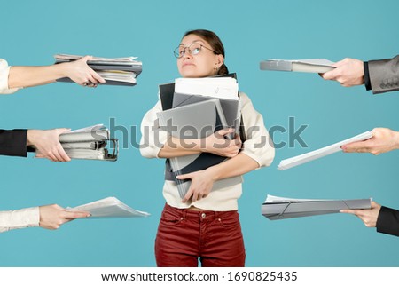 Girl hugs a large stack of documents and folders. At this time, employees are giving her a new job, loading the girl even harder