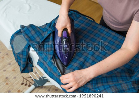 The girl is ironing a blue-checked shirt with a dark iron. Cleaning of the apartment.