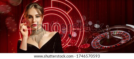 Girl in jewelry and black dress. Showing two red chips, posing on colorful background with neon lights, roulette and chips. Poker, casino. Close-up