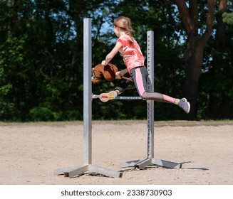 Girl jumping on hobby horse. Champion. Horse sport. Summer light. Green outdoor trees background. Child sport. Banner - Shutterstock ID 2338730903