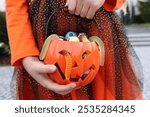 Girl kid in orange Halloween dress standing and holding in hands plastic pumpkin with assorted sweets. No face, unrecognizable. Halloween trick-or-treat concept.