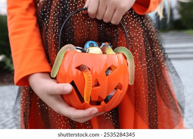 Girl kid in orange Halloween dress standing and holding in hands plastic pumpkin with assorted sweets. No face, unrecognizable. Halloween trick-or-treat concept. - Powered by Shutterstock