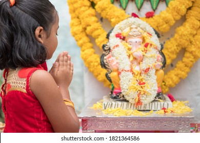 Girl Kid Praying By Closing Eyes And Folding Hands In Front Of Lord Ganesha Idol During Ganesha Or Vinayaka Chaturthi Festival Ceremony At Home - Concept Of Indian Religious Festival Celebrations