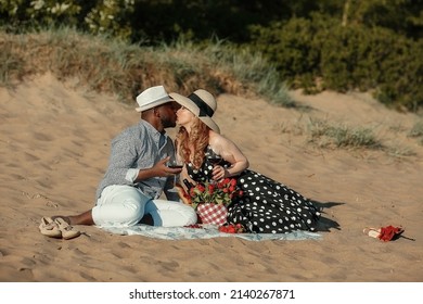 A Girl Kisses Her African American Boyfriend. A Young Interracial Couple Had A Picnic On The Beach. Man And Woman Drink Red Wine. Romantic Date On A Summer Day 