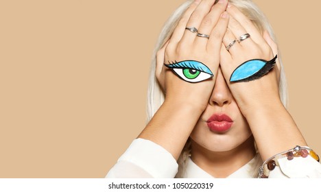 Girl kissing with closed and open eyes painted on her hands. Studio portrait of beautiful blonde model on beige background. Concept of flirting and fun.