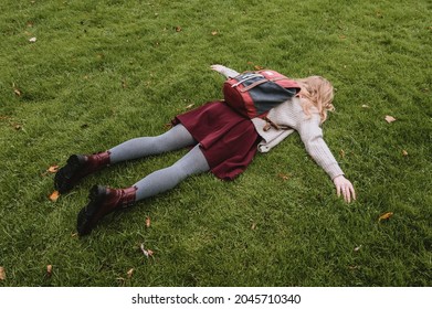 Girl in knitted jacket, burgundy skirt and leather shoes is lying face down on green grass in a city park. Tired of school.