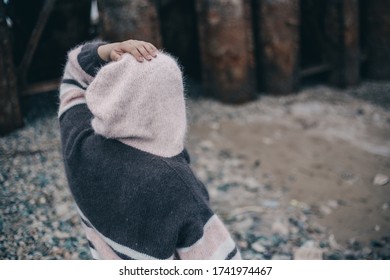 A Girl In A Knitted Jacket With A Hood Stands With Her Back Against The Background Of The River And Holds Her Head With Her Hand. Handmade Item Is Knitted With Merino Yarn Needles.