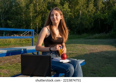 A girl with a laptop and books sits in the grandstand of the stadium, on the territory of the college. A healthy snack after classes. - Powered by Shutterstock