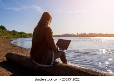 A Girl With A Laptop Is Sitting On A Log By The River Bank In A Park Outside The City. Concept Of Freelance, Remote Work Or Training. An Independent Female Businessman Works In Nature In Traveling.