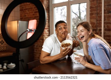 Girl Laughing From Guy Face In Sauce During Making Eat Fast Food Video Challenge On Smartphone. Cheerful Multiethnic Couple Of Generation Z Creating Content For Social Networks At Home. Video Blogging