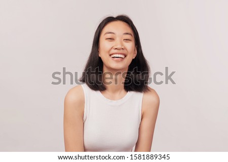 Girl laughs at something funny closes eyes with pleasure. Businesslike young woman Asian appearance with black hair and brown eyes dressed in short shirt stands isolated white background in Studio.