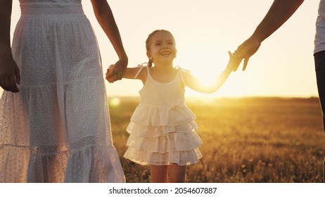 Girl Leads Her Mother And Father By The Hand At Sunset, A Little Daughter With Her Parents In The Sunshine, Hold Hand In Hand, A Happy Family, A Child With Mom And Dad, Take Care Of And Love The Baby