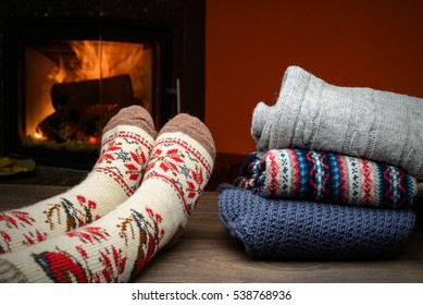 Girl Legs In Knitted Socks Near The Fireplace With A  Stack Of Cozy Knitted Sweaters On Old Table.