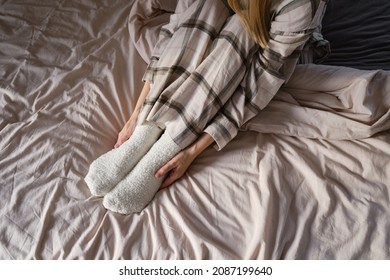 The Girl Lies In Bed In Pajamas And Warm Socks. Women's Legs In Cozy White Fluffy Socks In Bed. View From Above. Warm Evenings At Home