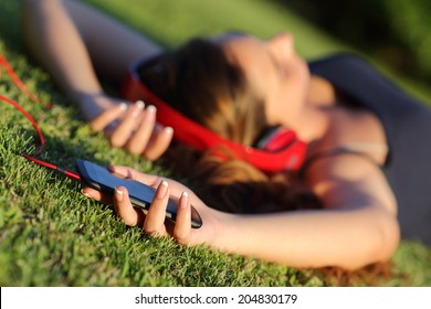 Girl Listening Music With Headphones And Holding A Smart Phone Lying On The Green Grass In A Park