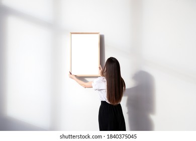 Girl with long black hair in white t-shirt standing right back and hanging light wooden mockup frame on white wall with shadow silhouettes