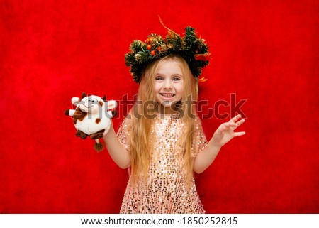 a girl with long blonde loose hair and a new year's wreath on her head with emotions on her face holds a soft toy in the form of a cow on a red background in her hand