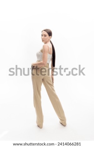A girl with long dark hair gathered in a ponytail, wearing beige trousers and a white top, stands half-turned against a white background.