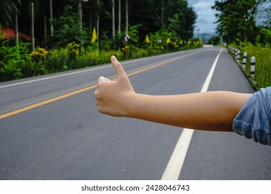 A girl with long hair, a gray long-sleeved shirt, and black pants is making hand signals to hitchhike on the side of the road.