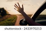 Girl with long hair sits in front seat of car, her hand out window and catching wind, glare of setting sun. Free woman driver rides car catches wind with her hand from car window. Child travels by car