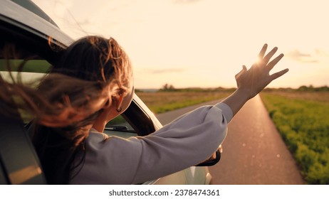 Girl with long hair sits in front seat of car, her hand out window and catching wind, glare of setting sun. Free woman driver rides car catches wind with her hand from car window. Child travels by car - Powered by Shutterstock