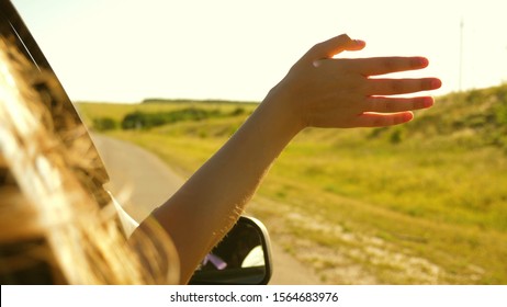 Girl With Long Hair Is Sitting In Front Seat Of Car, Stretching Her Arm Out Window And Catching Glare Of Setting Sun.