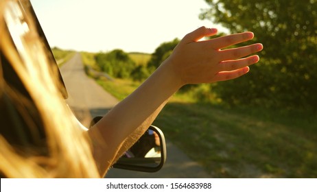 Girl With Long Hair Is Sitting In Front Seat Of Car, Stretching Her Arm Out Window And Catching Glare Of Setting Sun.