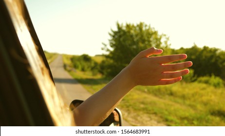 Girl With Long Hair Is Sitting In Front Seat Of Car, Stretching Her Arm Out Window And Catching Glare Of Setting Sun.