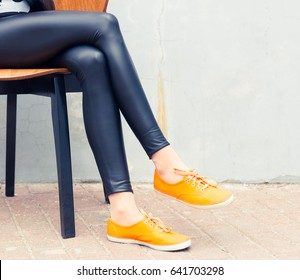 A girl with long legs in tight black pants and fashionable orange sneakers sits on a chair in the street. Outdoor. Fashion.