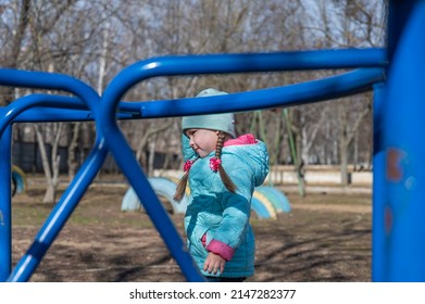 A Girl With A Long Pigtail Stands On The Playground. A Five-year-old Child Without A Care In The World. Blue Children's Swing In The Foreground.