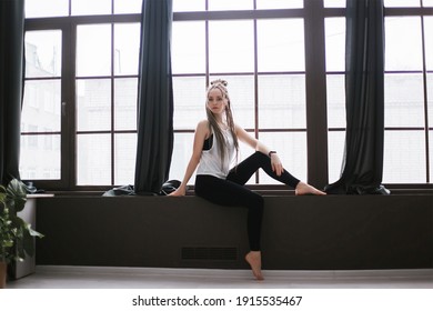 A girl with long pigtails sits on a windowsill near a large window. Relaxation while playing sports