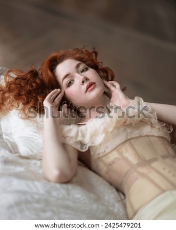A girl with long red hair in a lace chemise with a corset lies on the bed