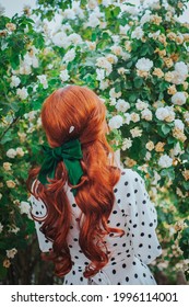 A Girl With Long Red Hair Stands Against The Backdrop Of A Blooming Garden. Back View. Women's Hairstyle With A Green Bow. Hair Ribbon.