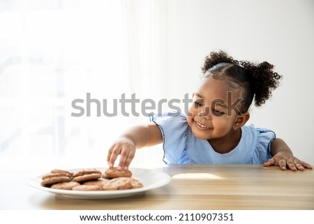 Girl looking at brown biscuit in the kitchen while her parents are cooking. Young girl pick up snacks on the table to secretly eat cookies.