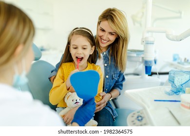 Girl looking in the mirror and brush her teeth after dental procedure while mother sitting near her for support. - Powered by Shutterstock