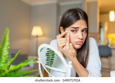 Girl looking at mirror and popping a pimple at home. Girl squeezing pimple at home. Woman examining her face in the mirror, problematic acne-prone skin concept. Upset teenager - Powered by Shutterstock